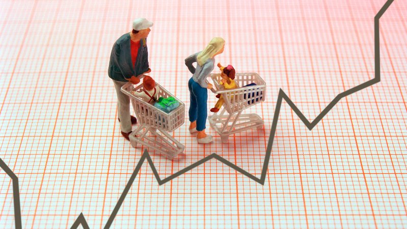 Eine Frau und ein Mann mit jeweils einem Kind in einem Einkaufswagen als Miniaturfiguren - als Untergrund ist ein Kurvendiagramm auf Millimeterpapier zu sehen. 
