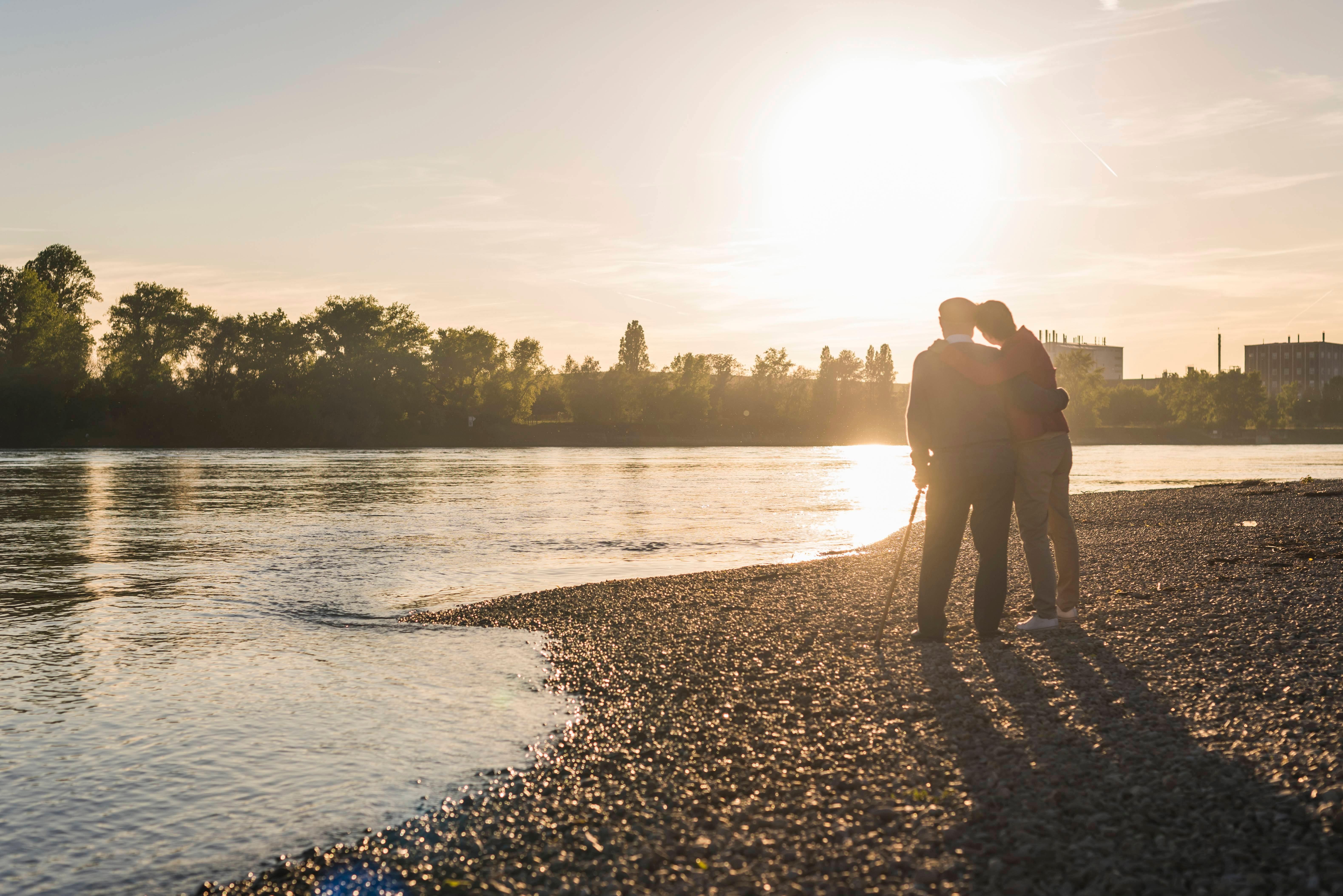 Ein älterer Mann steht mit seiner Tochter an einem Flusslauf und schaut auf den Sonnenuntergang (Archiv- und Symbolbild)