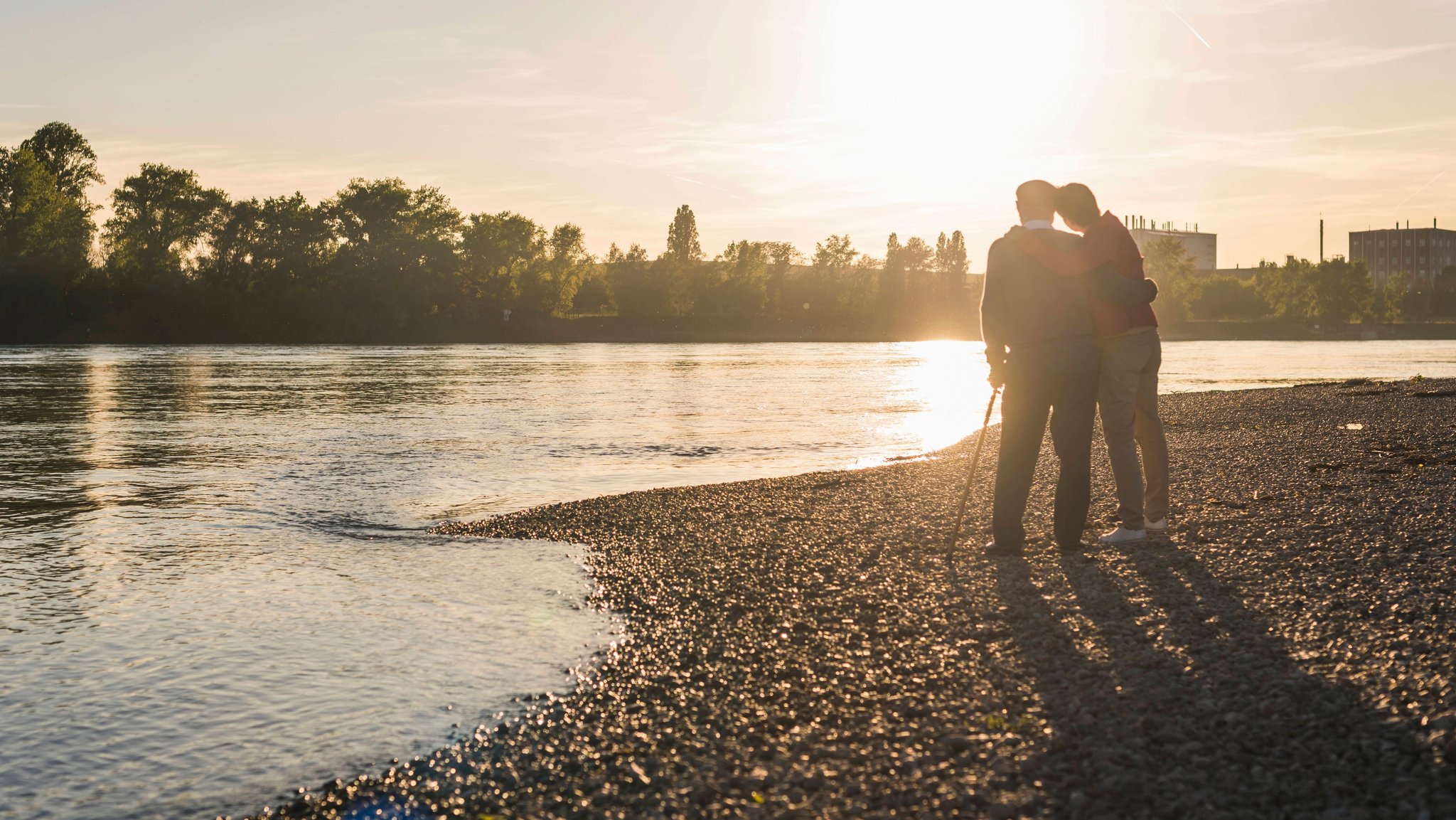 Ein älterer Mann steht mit seiner Tochter an einem Flusslauf und schaut auf den Sonnenuntergang (Archiv- und Symbolbild)