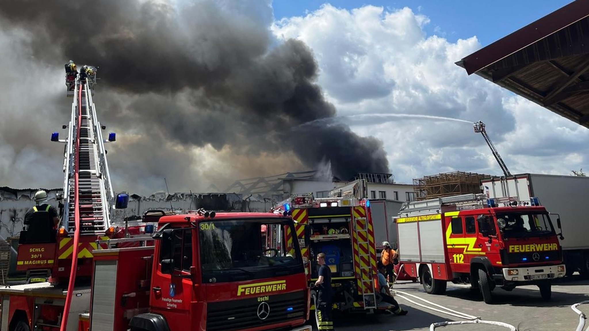 Feuerwehrkräfte versuchen den Brand in einer Lagerhalle zu löschen. Im Hintergrund ist eine schwarze Rauchsäule zu sehen.