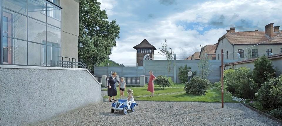 Zwei Frauen und zwei Kinder in einem sauberen Hof, im Hintergund der Wachtturm eines Konzentrationslagers. 