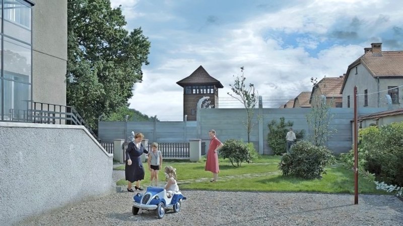 Zwei Frauen und zwei Kinder in einem sauberen Hof, im Hintergund der Wachtturm eines Konzentrationslagers. 