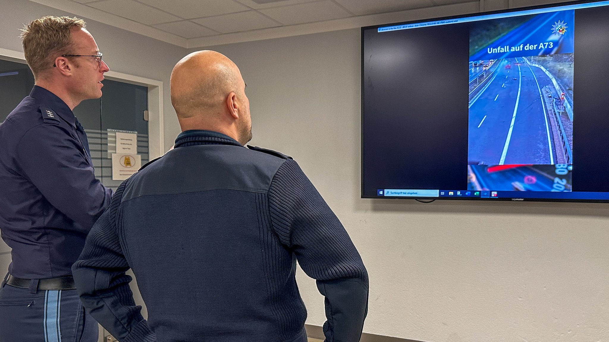 Zwei Polizisten stehen vor einem großen Bildschirm, auf dem ein Unfallvideo läuft.