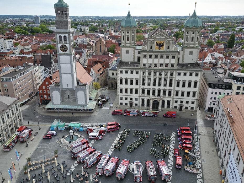 Vor dem Augsburger Rathaus haben sich Feuerwehrleute versammelt und bilden in Formation die Zahl "175". Um sie herum parken verschiedenste Feuerwehrfahrzeuge. Aufgenommen wurde das Foto anlässlich des 175-jährigen Jubiläums, das die Feuerwehr Augsburg am 14. und 15. September groß feiern wird.