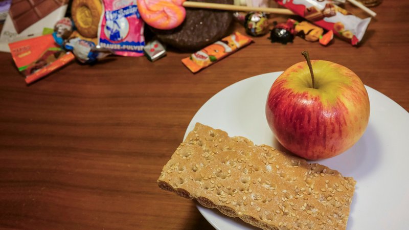 Apfel und Knäckebrot auf einem Teller neben Süßigkeiten. | Bild: picture alliance / epd-bild | Mika Preller-Pieroth Apfel und Knäckebrot auf einem Teller neben Süßigkeiten.