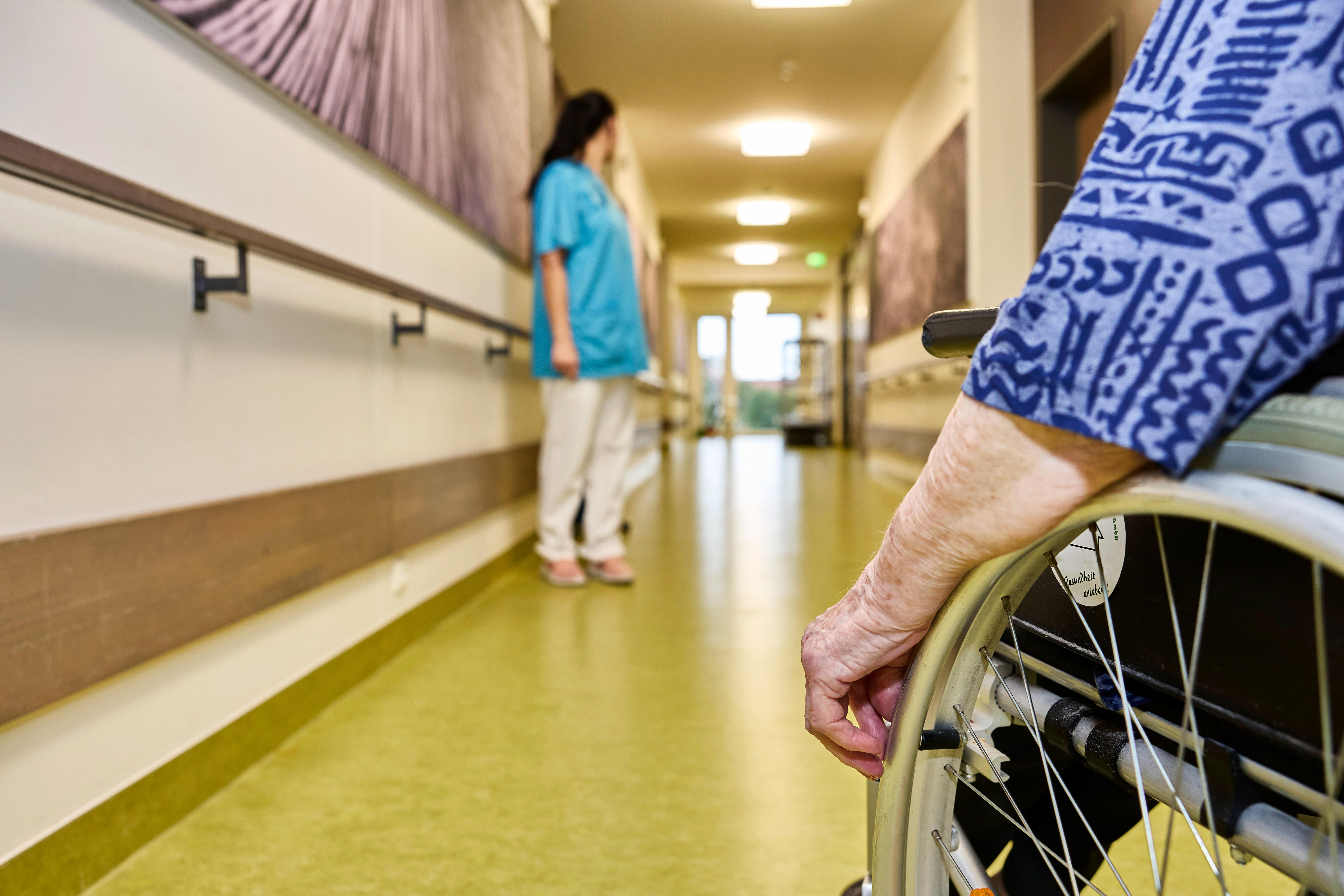 Symbolbild: Pfllegekraft und Bewohnerin im Rollstuhl auf dem Flur in einem Pflegeheim. 