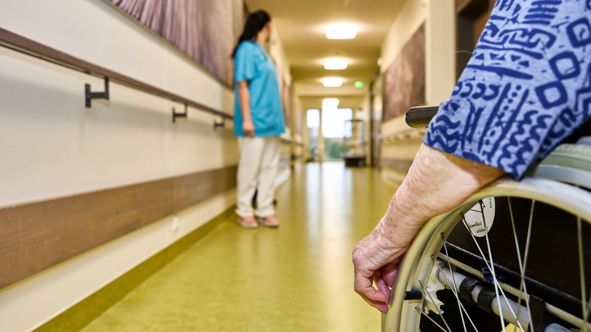 Symbolbild: Pfllegekraft und Bewohnerin im Rollstuhl auf dem Flur in einem Pflegeheim. 