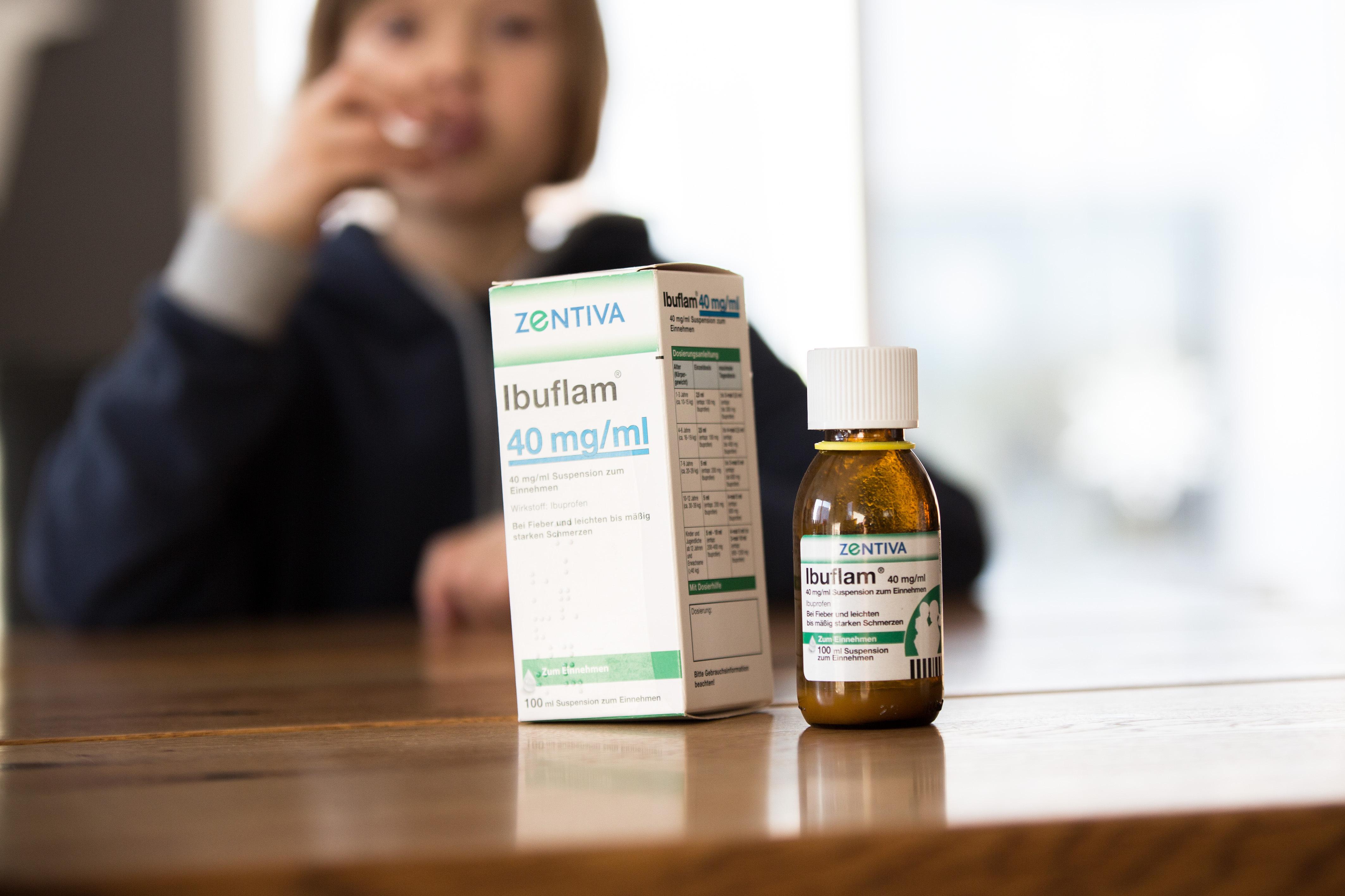 Auf einem Tisch stehen Fieber- und Schmerzmittelsaft für Kinder, im Hintergrund sitzt ein krankes Kind das den Schmerzsaft gegen Fieber sowie Schmerzen einnimmt. 