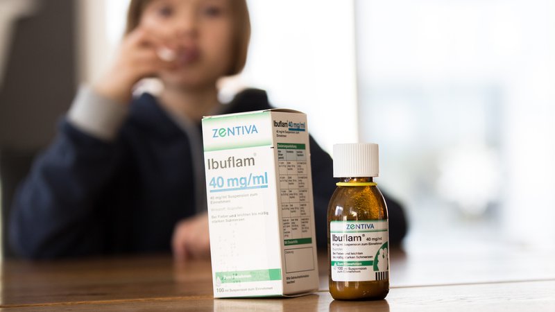 Auf einem Tisch stehen Fieber- und Schmerzmittelsaft für Kinder, im Hintergrund sitzt ein krankes Kind das den Schmerzsaft gegen Fieber sowie Schmerzen einnimmt. | Bild: dpa/Fotostand/K. Schmitt Auf einem Tisch stehen Fieber- und Schmerzmittelsaft für Kinder, im Hintergrund sitzt ein krankes Kind das den Schmerzsaft gegen Fieber sowie Schmerzen einnimmt.