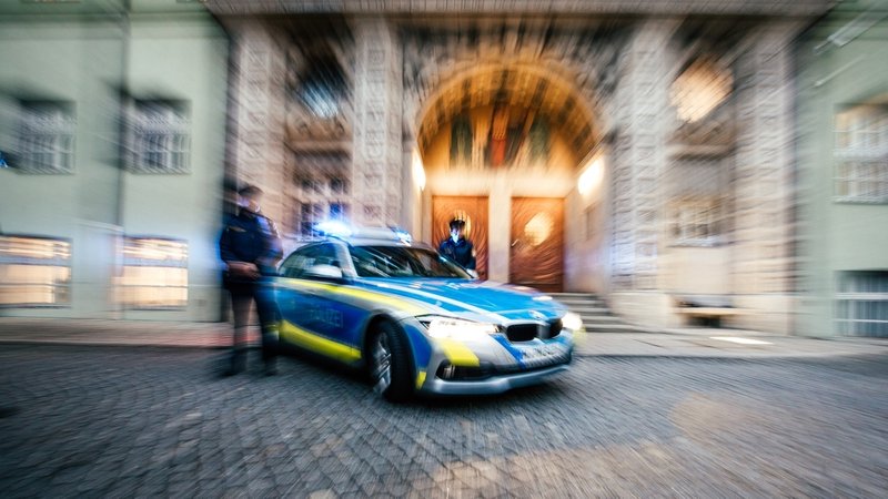 Eine Polizistin und ein Polizist stehen vor dem Eingang des Polizeipräsidiums München vor einem Polizeiauto mit Blaulicht. (Symbolbild)