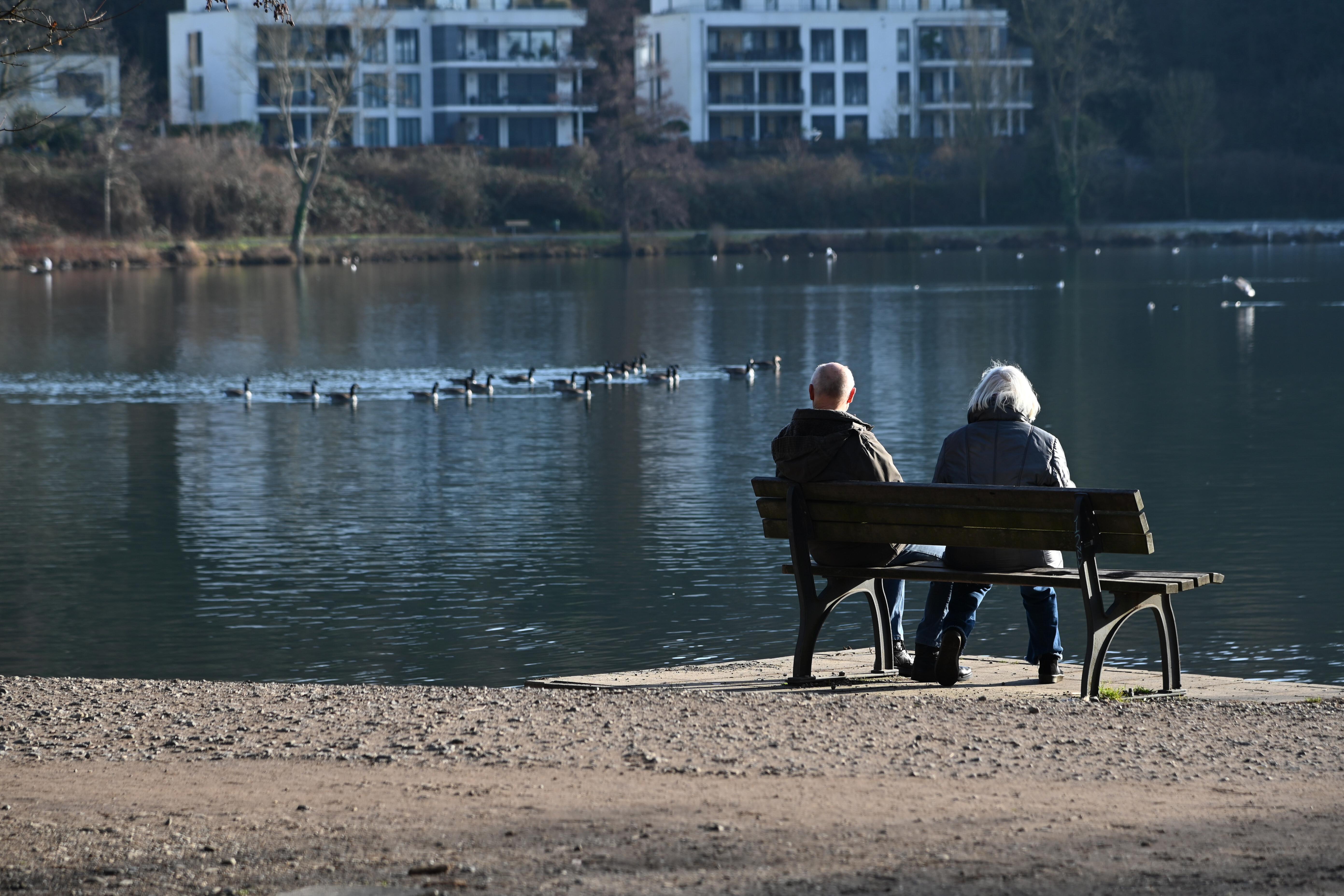 Zwei Senioren sitzen auf einer Bank und schauen auf einen See mit Gänsen.
