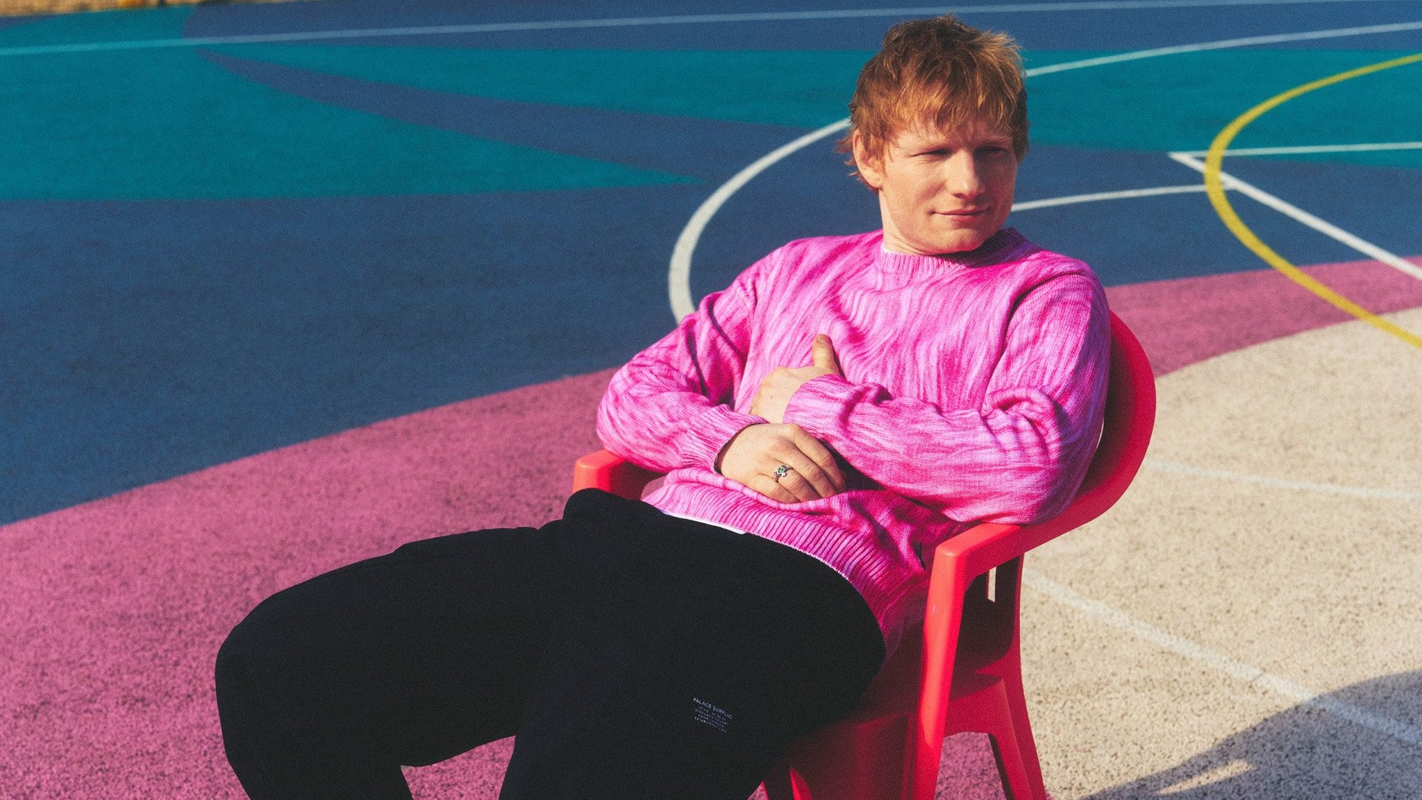 Ed Sheeran sitzt mit einem pinken Pullover auf einem leeren Sportplatz in einem roten Plastikstuhl
