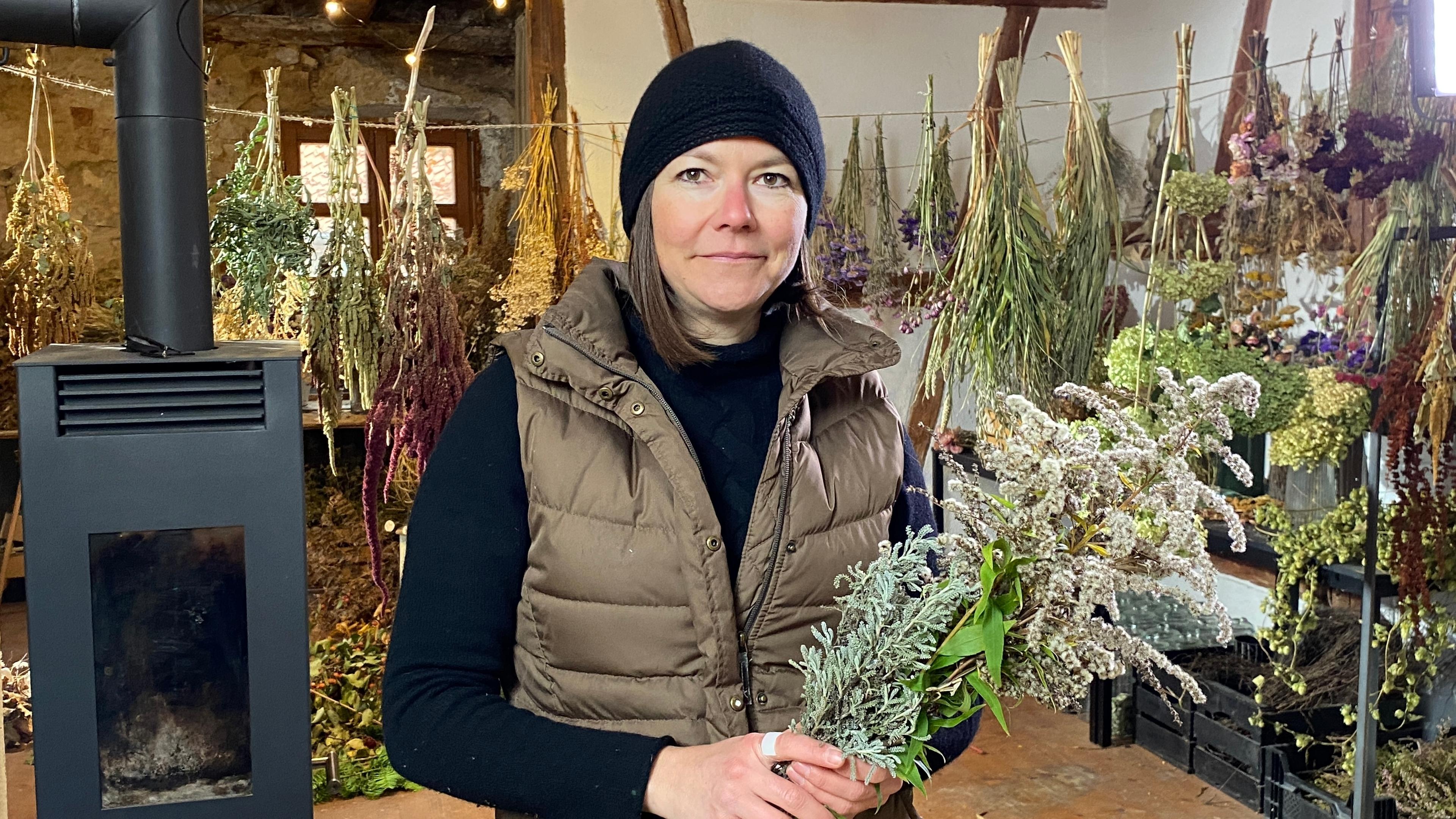 Drei Ideen für natürliche Adventsdeko. Anja Schickaneder (Neunhof, Lkr. Nürnberger Land) steht mit einem Strauß Trockenblumen im Arm in einem Raum mit Schwedenofen. An der Wand hängen verschiedenste Trockenblumen und getrocknete Naturmaterialien. 