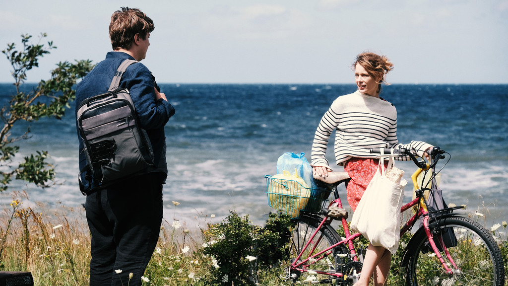 Leon in schlabbrigen Klamotten und mit Rucksack über der Schulter trifft am Ostsee-Ufer auf Nadja, die mit einem Fahrrad unterwegs ist.
