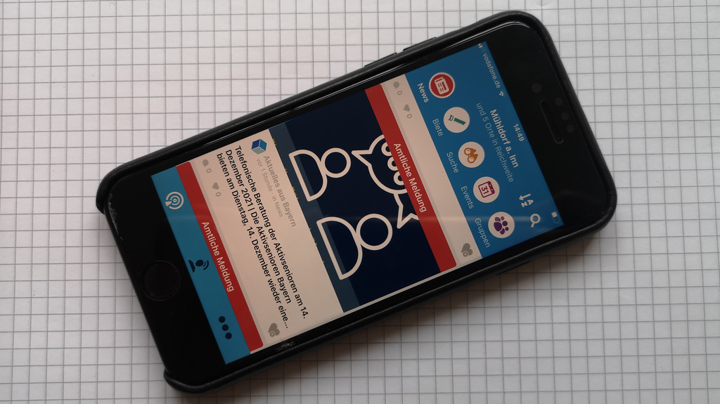 Auf dem Smartphone sieht man die Oberfläche der BayernFunk-App mit den Reitern News, Biete, Suche, Events und Gruppen. Darunter sieht man eine amtliche Meldung.
