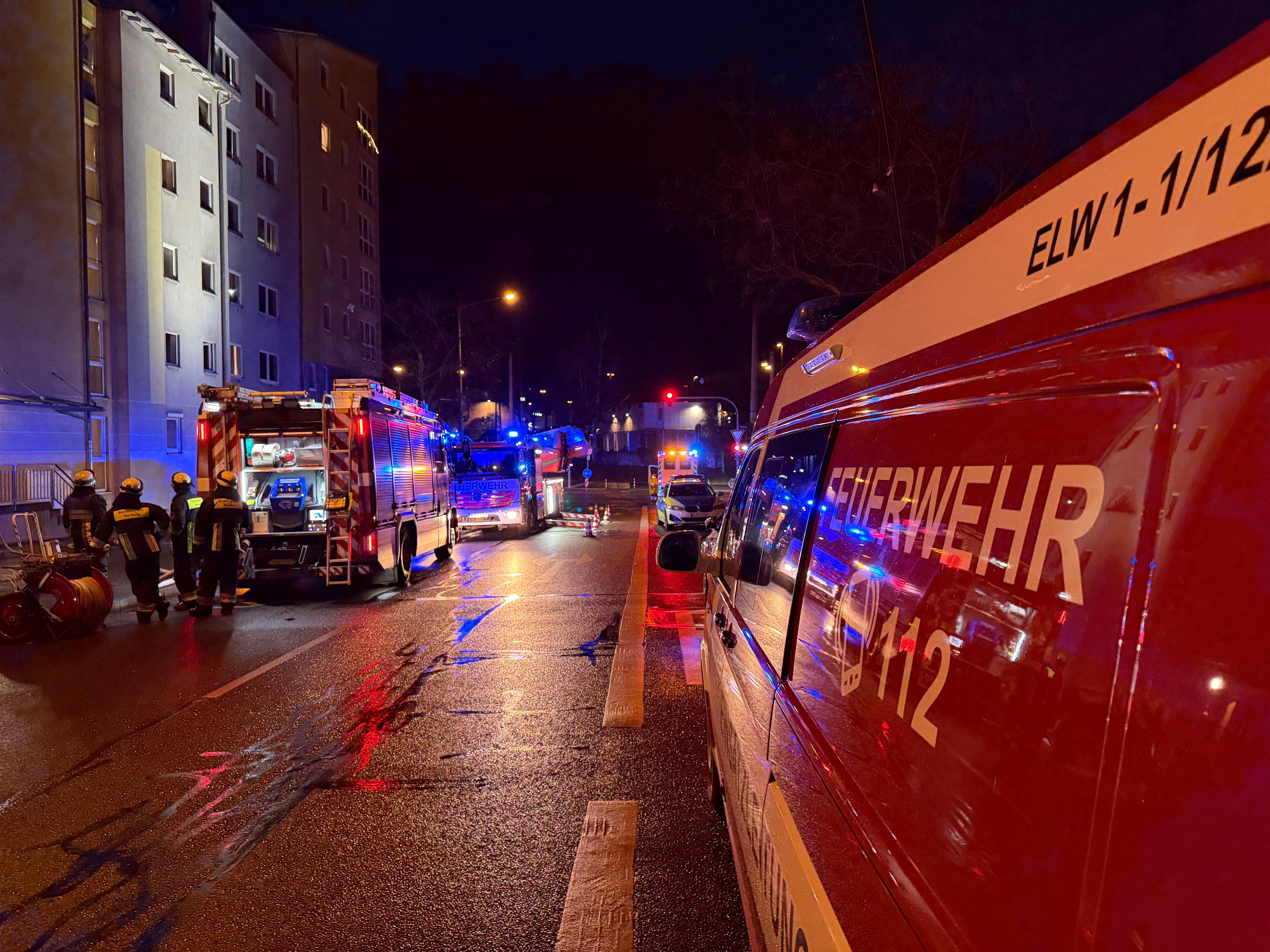Einsatzfahrzeuge der Feuerwehr stehen auf einer regennassen Straße vor einem Mehrfamilienhaus. Auf der Straße sind mehrere Einsatzkräfte zu sehen. 