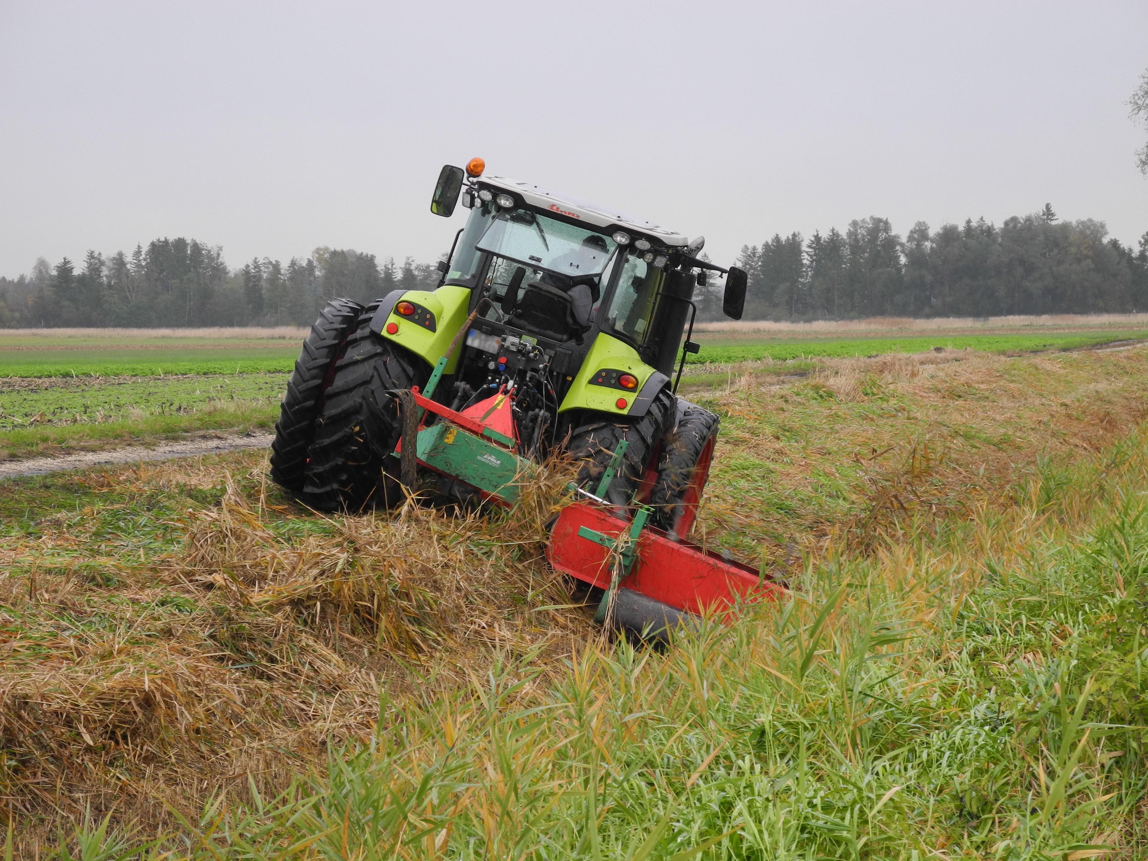 Ein hellgrüner Traktor mit Doppelbereifung von hinten. Er fährt eine Grabenböschung entlang und hat hinten einen grünroten Bandrechen angebaut
