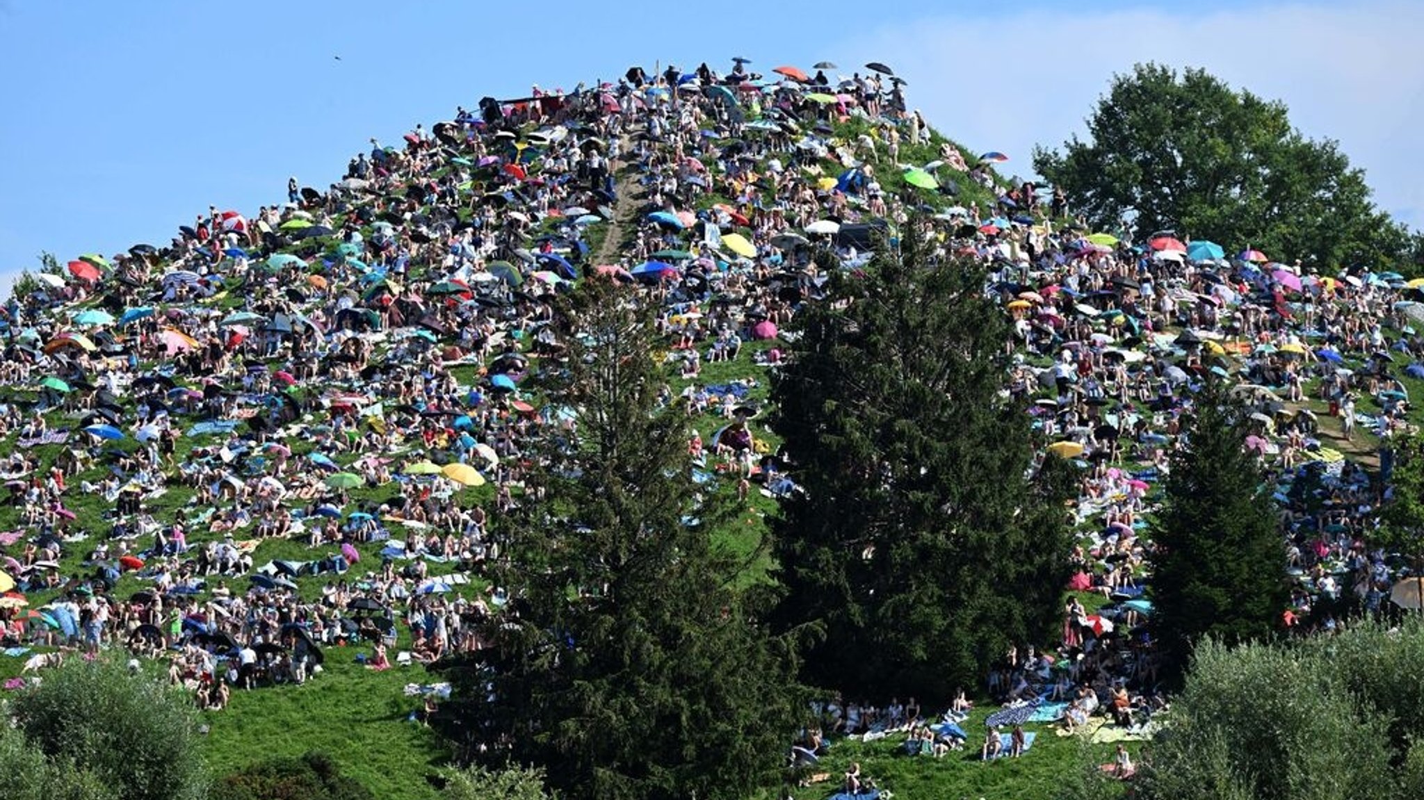 Fans sitzen auf dem Olympiaberg im Olympiapark und warten bei Sonnenschein auf den Beginn des Taylor Swift Konzerts