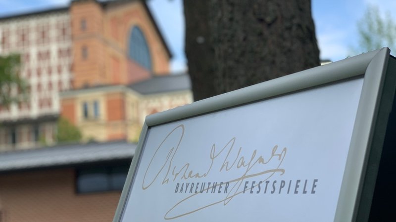Auf einem Schild steht "Richard Wagner Bayreuther Festspiele", im Hintergrund ist das Festspielhaus zu sehen. 