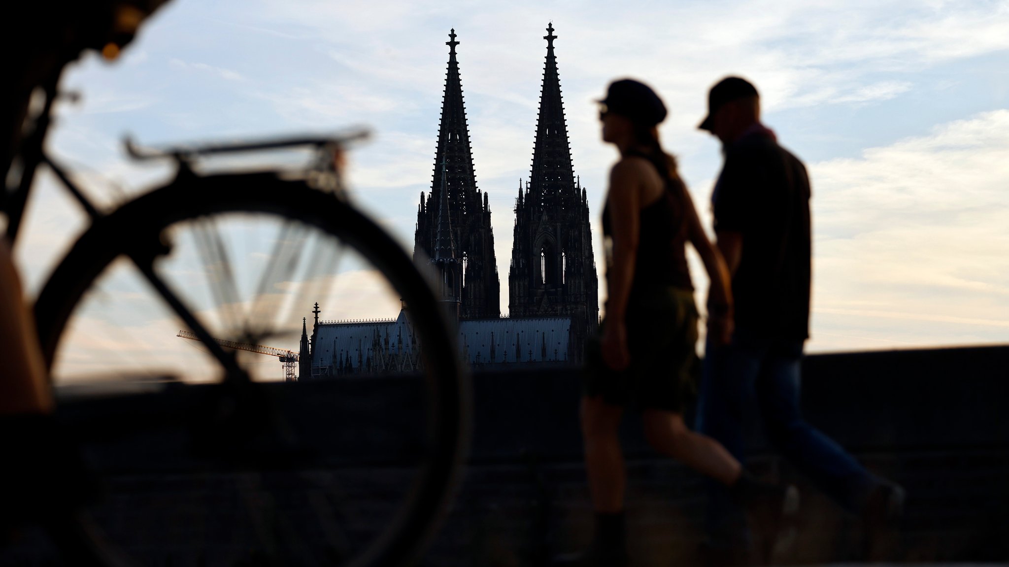 Passanten am Kölner Rheinboulevard, im Hintergrund ragt die Doppelspitze des Kölner Dom in den Abendhimmel. 