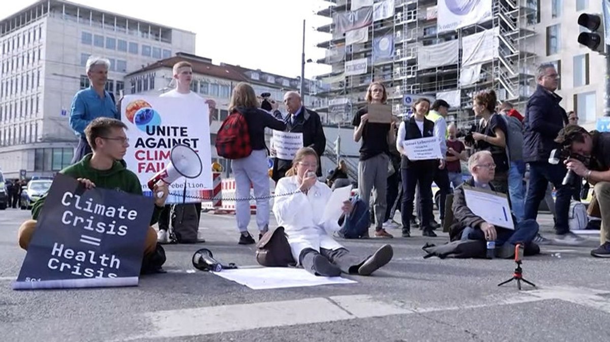 Klimaaktivisten sitzen und stehen auf der Straße, halten Schilder hoch, eine Frau spricht in ein Megafon.