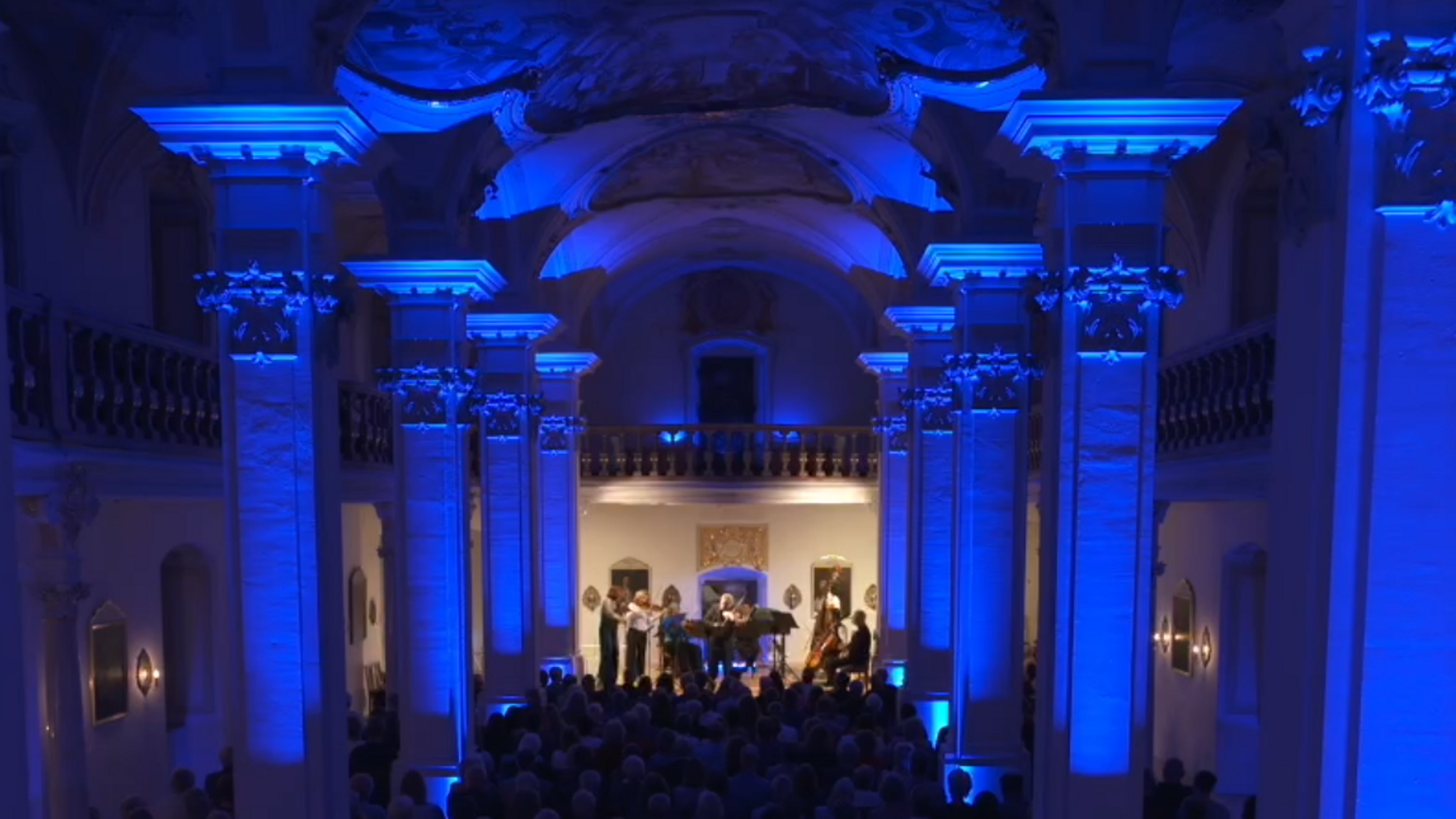 Ein Musikensemble spielt in einem Saal mit blau angeleuchteten Säulen und Deckenfresken.