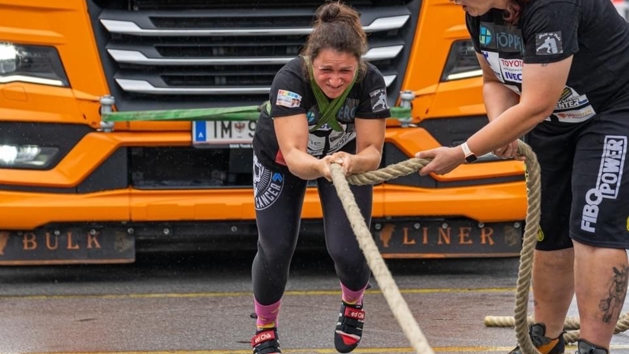 Eine Frau ist mit zwei vor der Brust kreuzenden Bändern vor einen LKW geschnürt und zieht diesen, indem sie sich an einem Seil nach vorne hangelt. Ihr Gesicht ist angestrengt verzogen. 