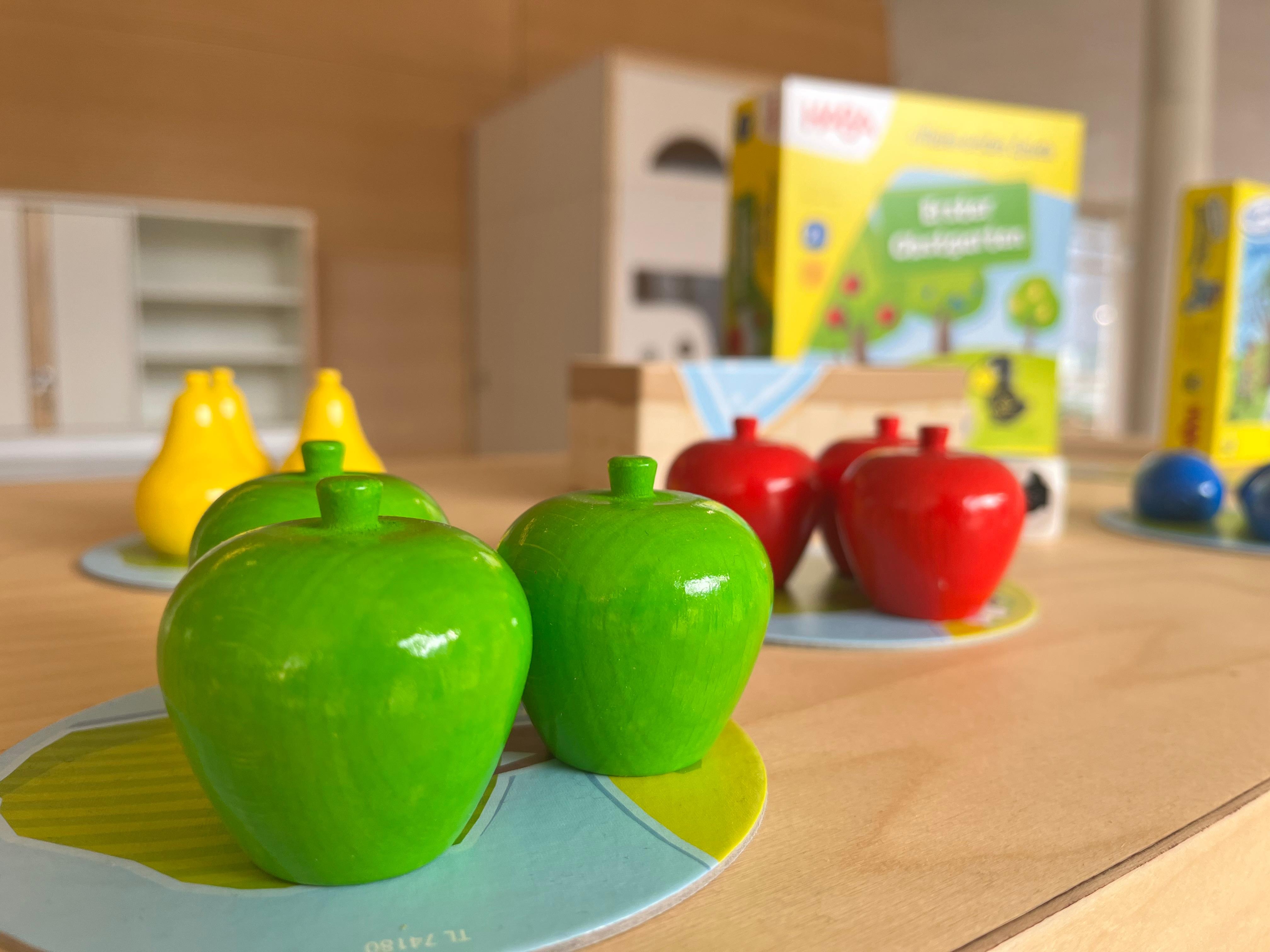 Holzobst steht auf einem Tisch. Im Hintergrund ist die Verpackung des Spiels "Obstgarten" von Haba zu sehen.