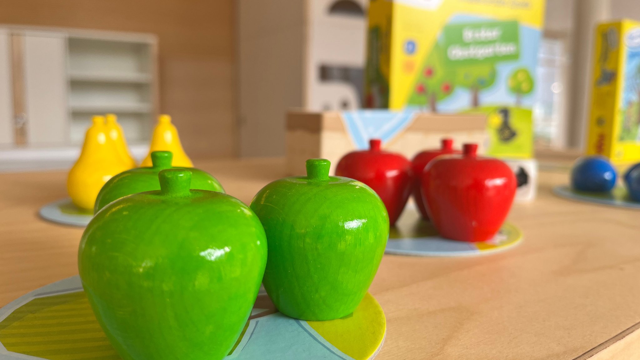 Holzobst steht auf einem Tisch. Im Hintergrund ist die Verpackung des Spiels "Obstgarten" von Haba zu sehen.