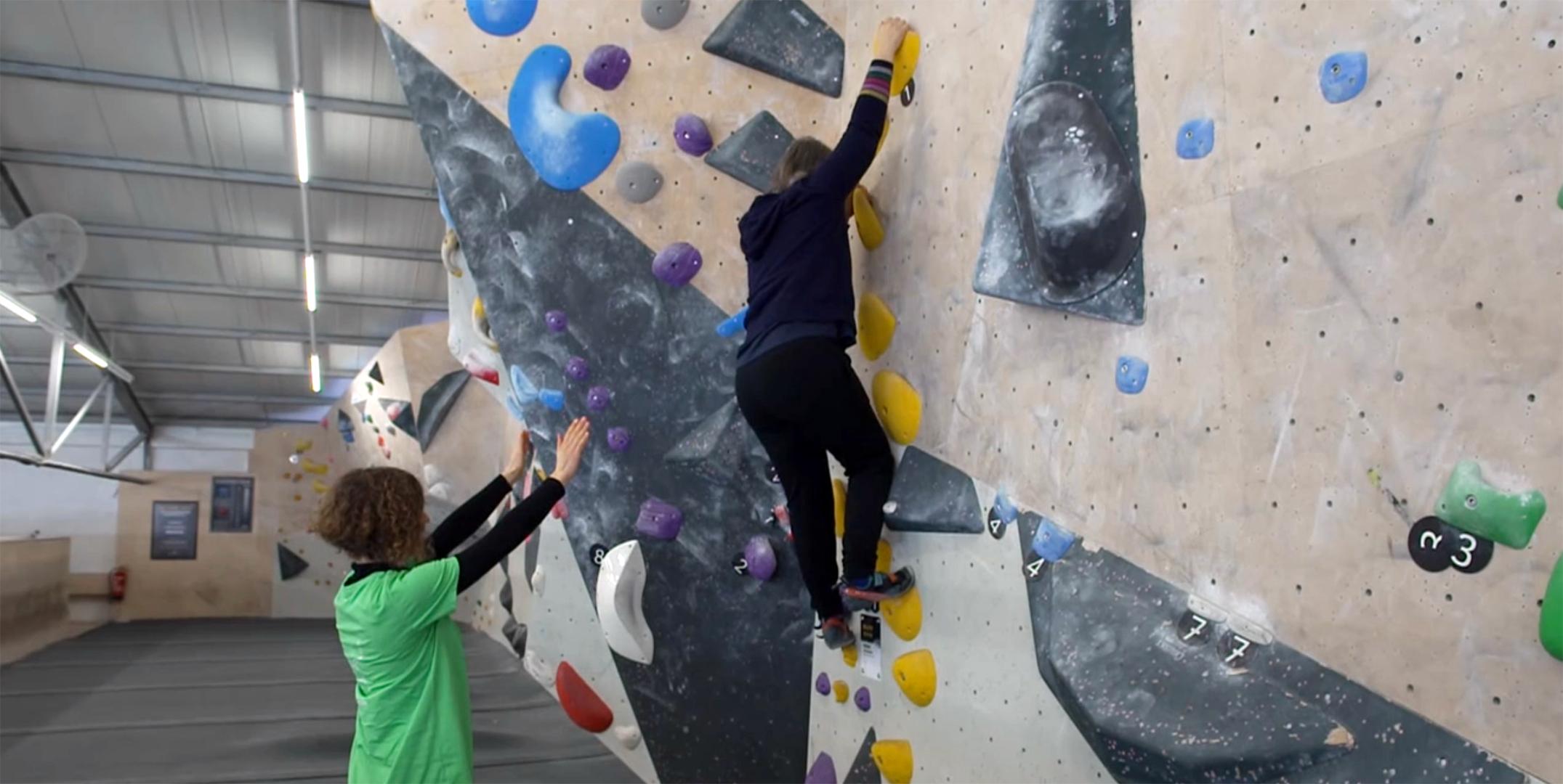Eine junge Frau an der Boulderwand, hinter ihr breitet eine Frau am Boden stehend schützend ihre Arme aus