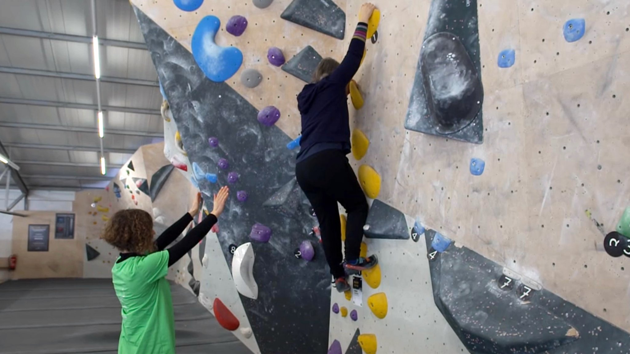 Eine junge Frau an der Boulderwand, hinter ihr breitet eine Frau am Boden stehend schützend ihre Arme aus