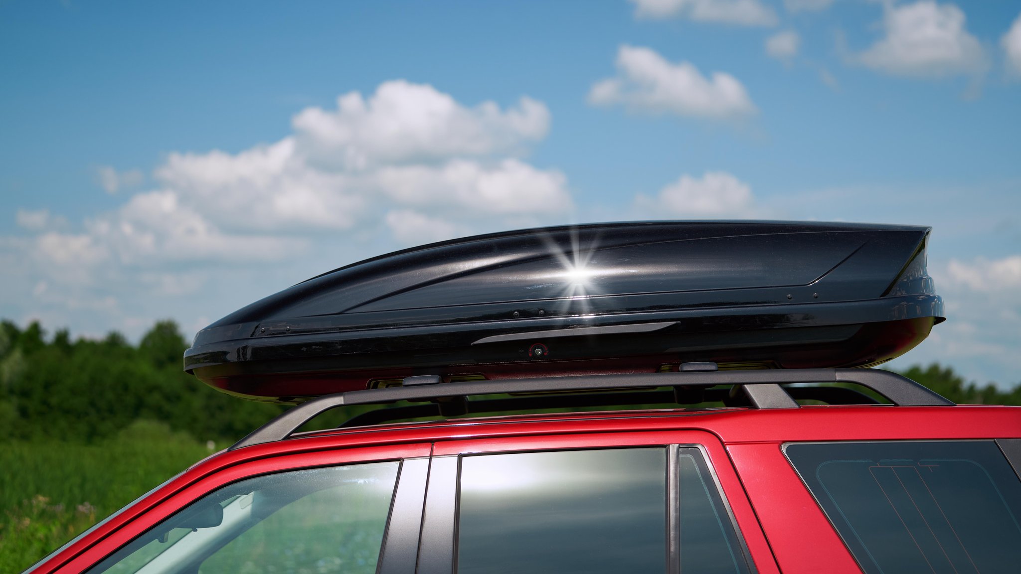 Nahaufnahme einer schwarzen glänzenden Dachbox auf einem roten Auto. Im Hintergrund ist ein grüner Wald und ein blauer Himmel mit Wolken