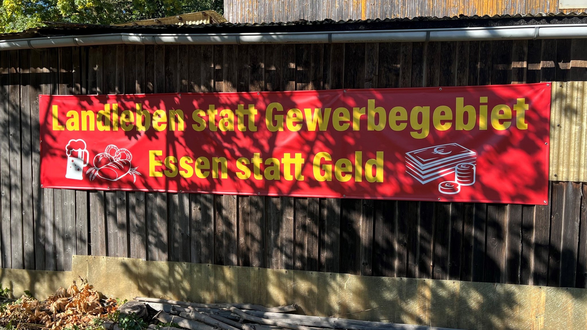 Ein rotes Protestplakat hängt an einer einer Scheunenwand, darauf ist in gelben Buchstaben die Aufschrift "Landleben statt Gewerbegebiet - Essen statt Geld" zu lesen.