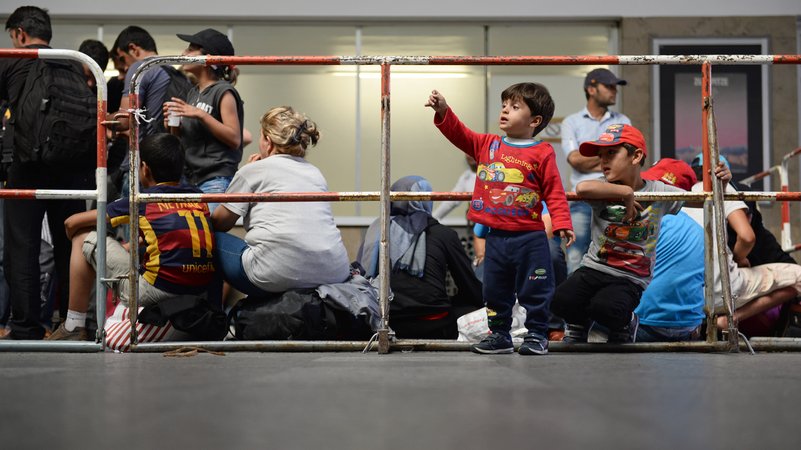 Flüchtlinge aus Syrien, die auf ihre Registrierung warten, stehen und sitzen am 31.08.2015 hinter einem Absperrgitter im Hauptbahnhof in München. | Bild: pa/dpa/Andreas Gebert Flüchtlinge aus Syrien, die auf ihre Registrierung warten, stehen und sitzen am 31.08.2015 hinter einem Absperrgitter im Hauptbahnhof in München.