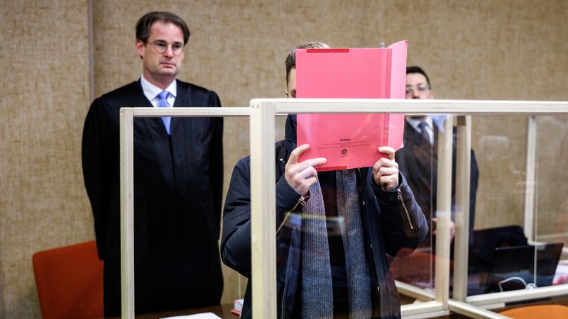Der Angeklagte (Mitte) steht am 15.2.23 im Gerichtssaal vor seinem Rechtsanwalt Florian Schmidtke (l) und verdeckt sein Gesicht.