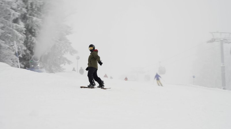 Snowboardfahrer auf einer Skipiste. Im Hintergrund bläst eine Schneekanone Kunstschnee in die Luft und die Gondeln der Bergbahn verschwinden im Nebel. | Bild: BR/Johannes Hofmann Snowboardfahrer auf einer Skipiste. Im Hintergrund bläst eine Schneekanone Kunstschnee in die Luft und die Gondeln der Bergbahn verschwinden im Nebel.