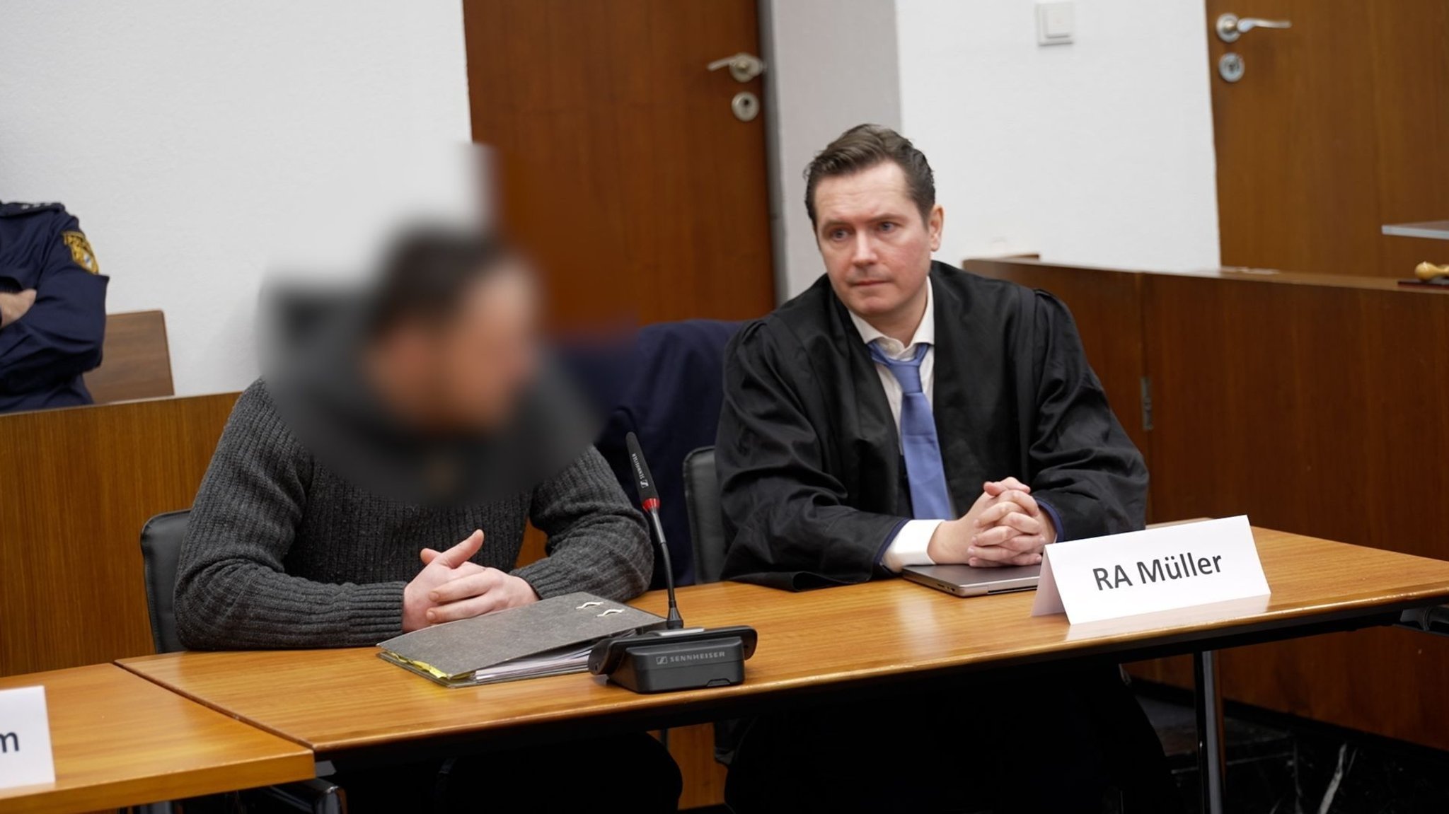 Der Angeklagte sitzt neben seinem Rechtsanwalt auf der Anklagebank. Vor dem Anwalt steht ein Schild mit der Aufschrift "RA Müller". Der Angeklagte ist verpixelt. 