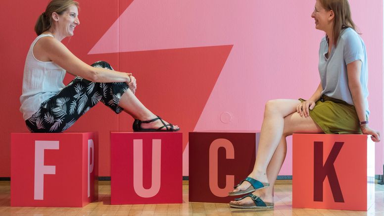 Heiter und gelassen: Zwei Damen in der Schimpf-Ausstellung - auf den Buchstabenwürfel, auf denen sie sitzen, steht F-U-C-K | Bild:Museum für Kommunikation Nürnberg