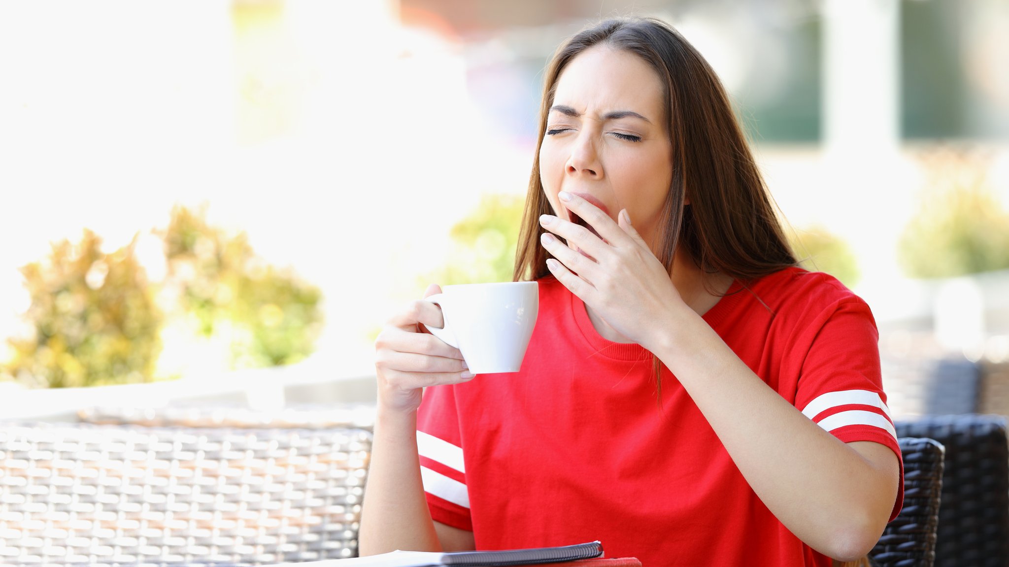 Frau im roten T-Shirt sitzt am Tisch, gähnt und hält eine Tasse Kaffee in der Hand