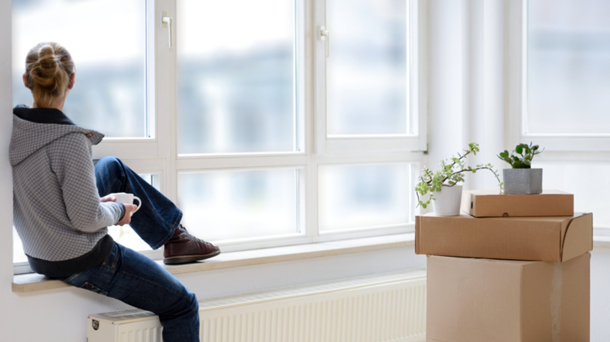 (Symbolbild) Eine junge Frau sitzt auf einer Fensterbank und schaut raus, neben ihr stehen Umzugskartons.