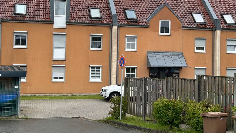 Ein braunes Mehrfamilienhaus. Im Vordergrund steht ein weißes Auto. Am Rand des Parkplatzes ist eine Biotonne und das Verkehrszeichen "absolutes Halteverbot" zu sehen. | Bild:BR/Markus Feulner