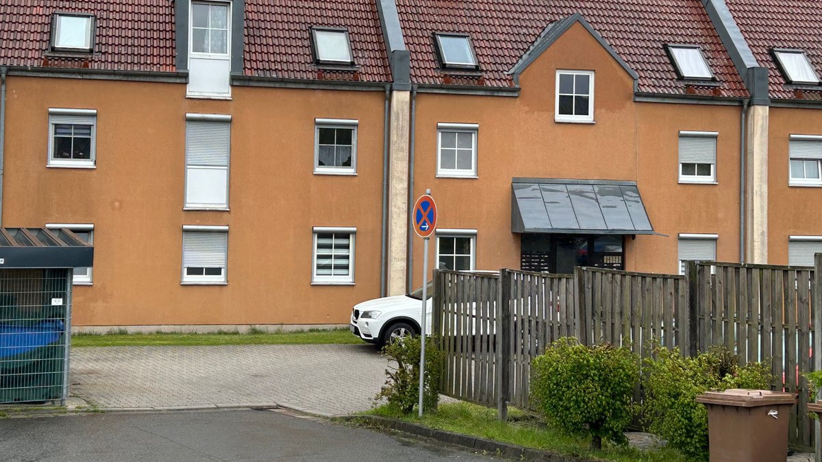 Ein braunes Mehrfamilienhaus. Im Vordergrund steht ein weißes Auto. Am Rand des Parkplatzes ist eine Biotonne und das Verkehrszeichen "absolutes Halteverbot" zu sehen.