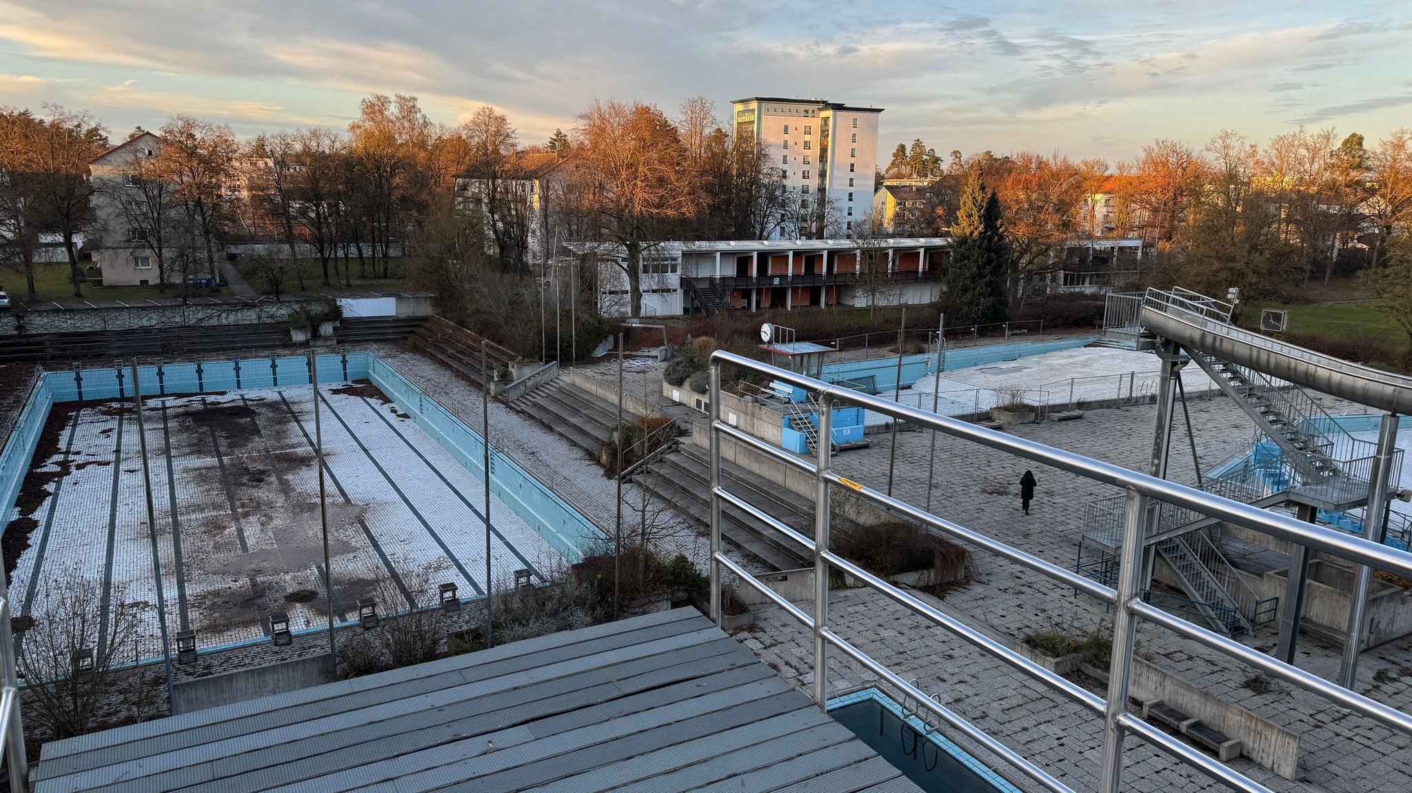 Von einem zehn Meter hohen Sprungturm aus sieht man ein großes, leeres, verdrecktes Schwimmbecken. Außerdem sind noch zwei weitere, leere Becken, eine Wasserrutsche und ein Bademeisterhäuschen zu sehen.