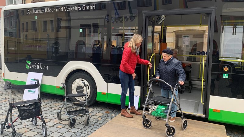 Eine Trainerin zeigt einer älteren Dame mit Rollator, wie sie aus dem Bus aussteigen kann.