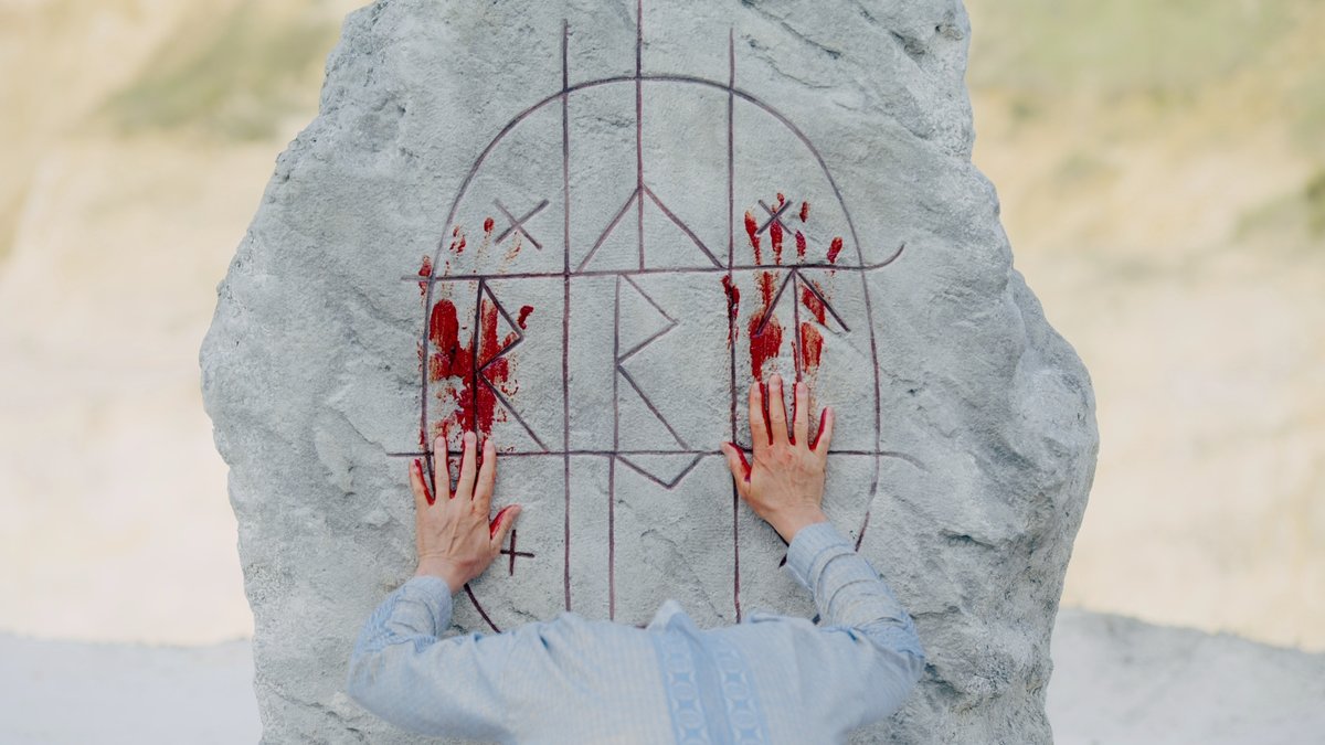 Eine Person mit abgesenktem Kopf und blutigen Händen hinterlässt eine tiefrote Spur auf einem weißen Felsen, der mit großen Runen versehen ist