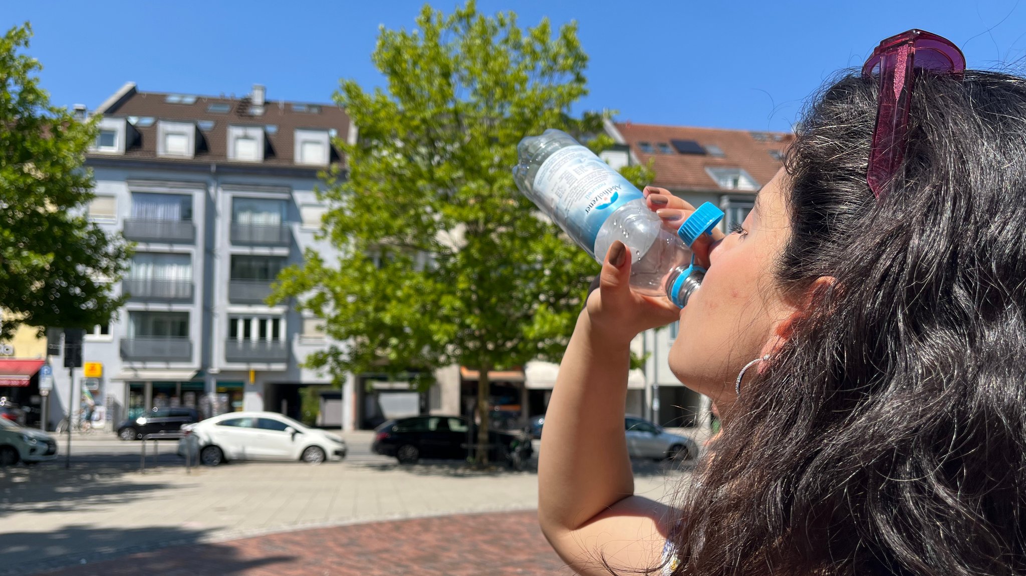 Eine Frau steht in der Stadt auf einem Platz in der Sonne und trinkt aus einer Wasserflasche. | Bild: BR/Rebecca Reinhard Eine Frau steht in der Stadt auf einem Platz in der Sonne und trinkt aus einer Wasserflasche.