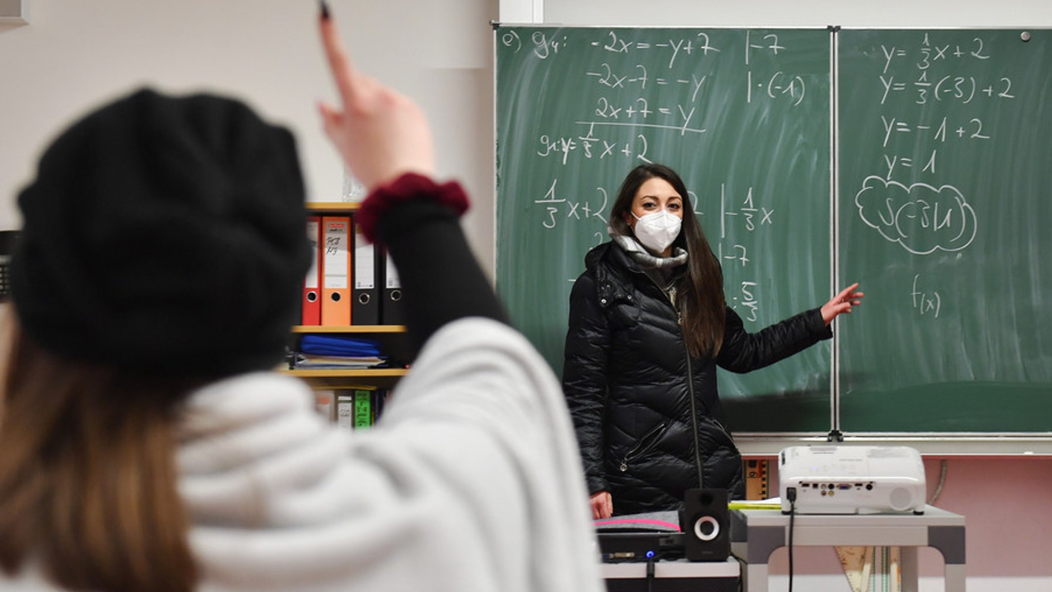 Lehrerin mit FFP2-Maske und Winterjacke unterrichtet in einem Klassenzimmer