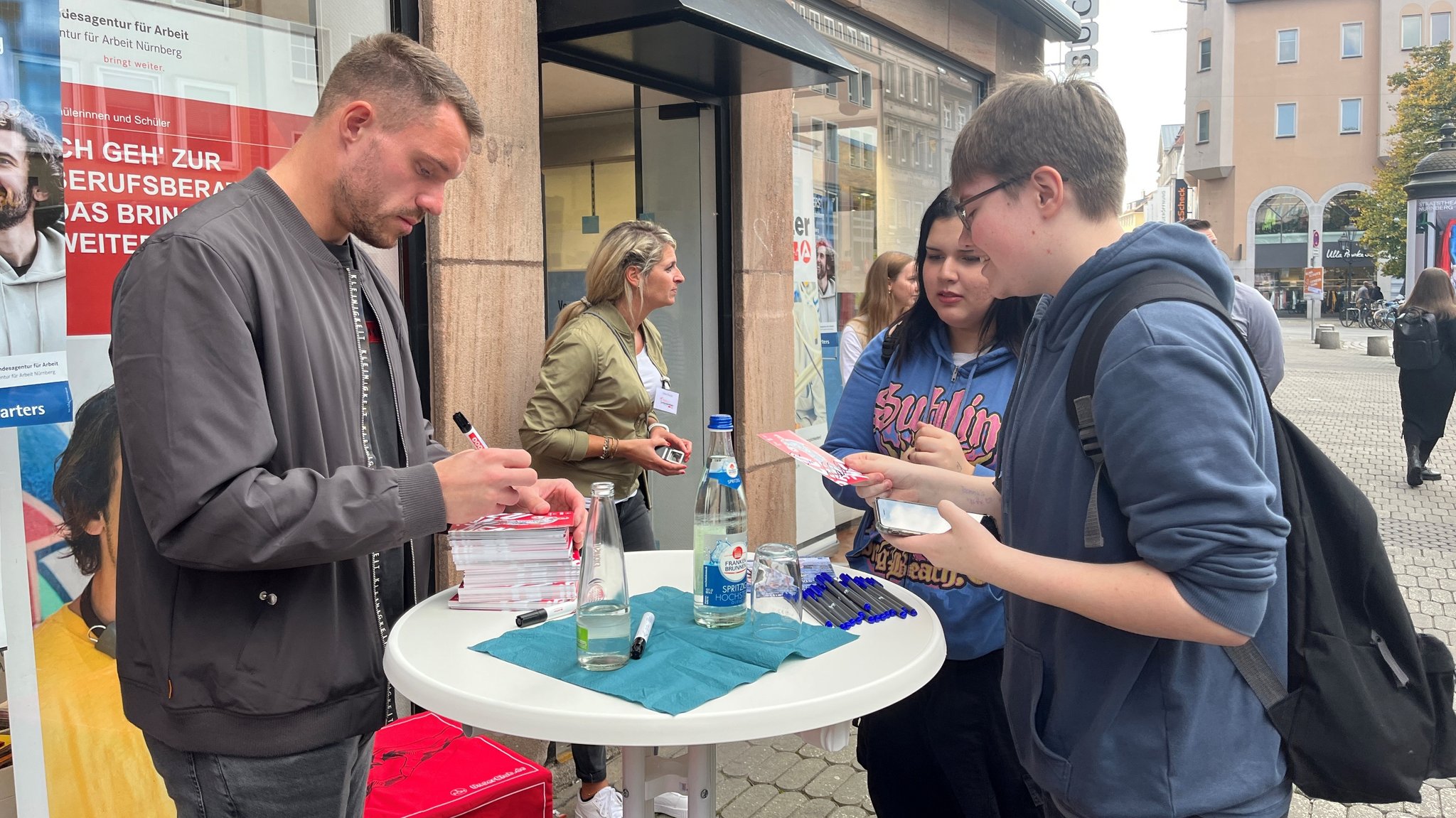 Ein Mann steht vor einem Laden an einem Stehtisch und schreibt Autogramme für zwei Jugendliche.