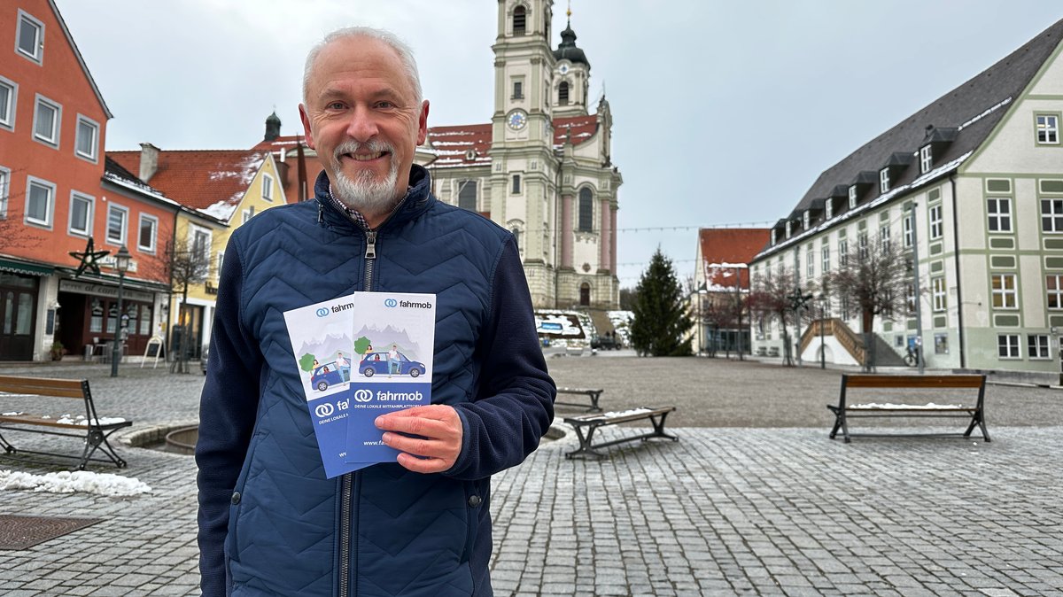 Helmut Scharpf steht vor der prächtigen Klosterkirche am Marktplatz in Ottobeuren und hält zwei Flyer in die Kamera