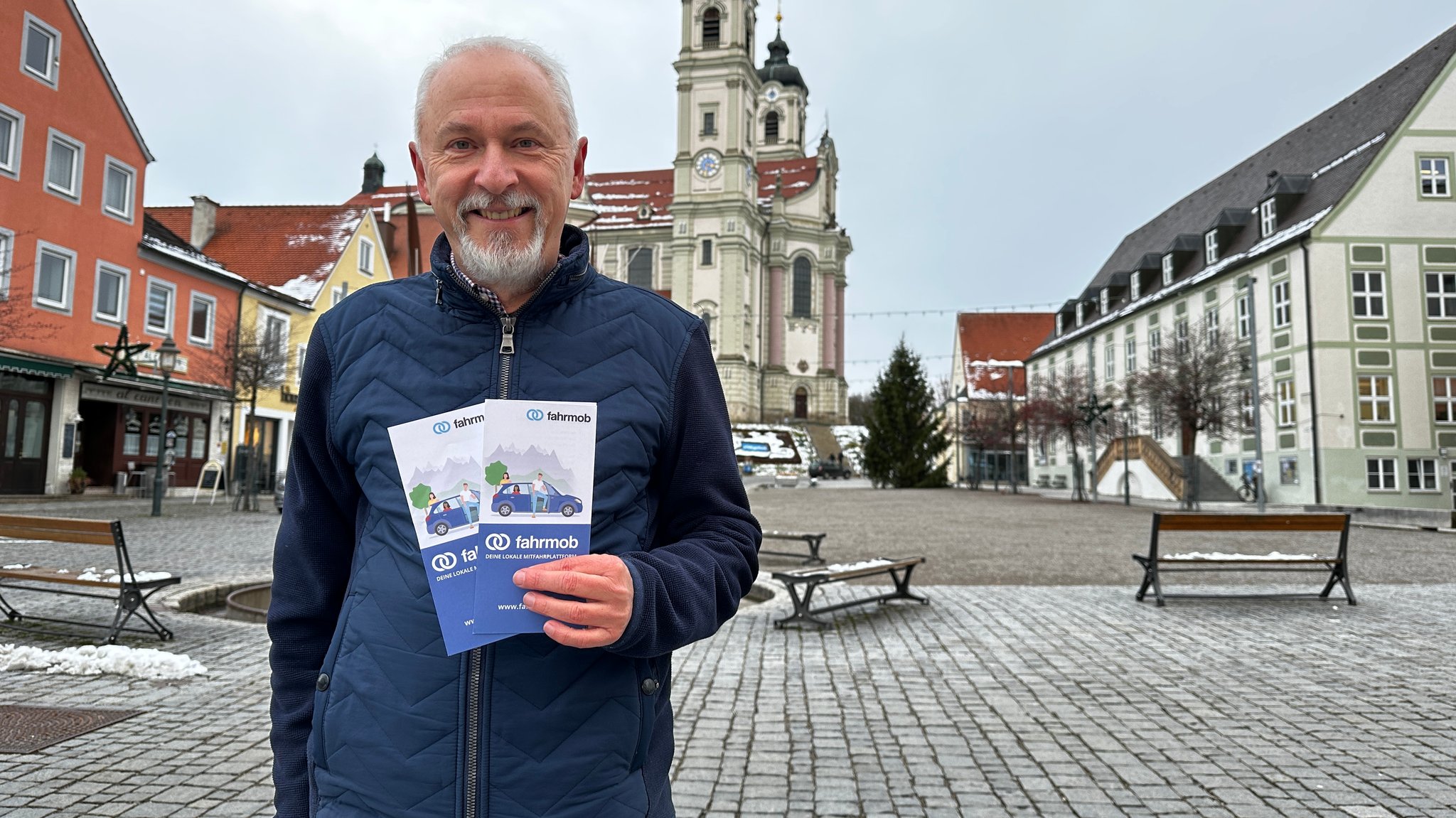 Helmut Scharpf steht vor der prächtigen Klosterkirche am Marktplatz in Ottobeuren und hält zwei Flyer in die Kamera